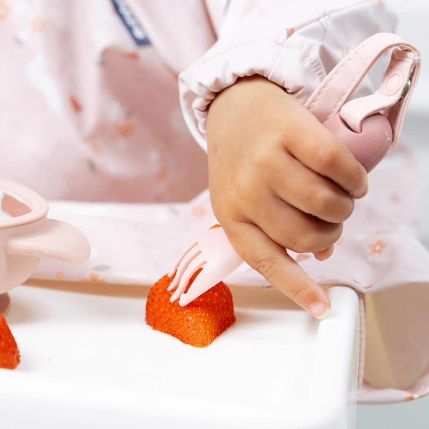 Toddler hand holding attached training fork to pick up a strawberry piece on a highchair tray, showing supported self-feeding in action.