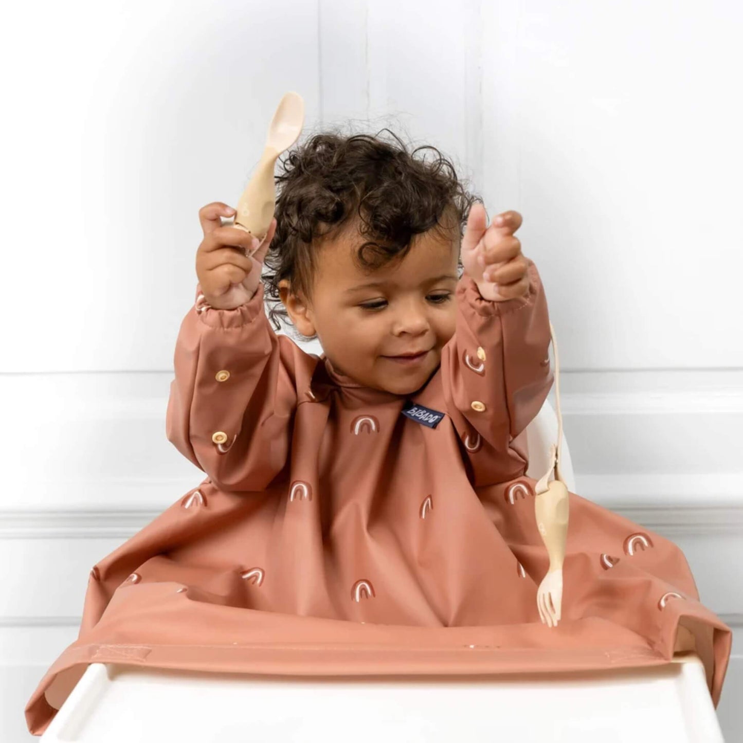 A toddler seated in a highchair wearing a rust rainbow long sleeve coverall bib while holding attachable cutlery during self-feeding.