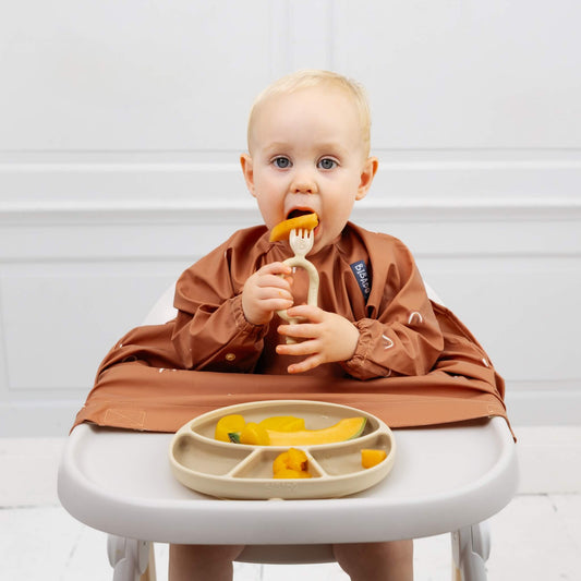 A baby using a fawn self-feeding utensil to eat melon while sitting in a highchair wearing a Bibado coverall bib.