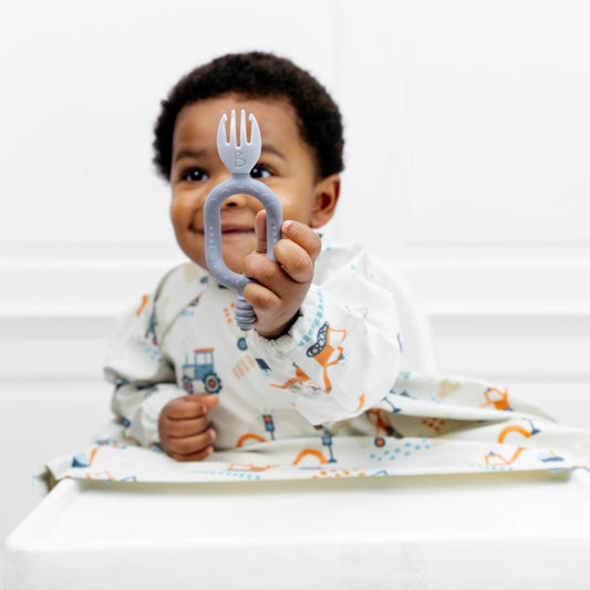 A smiling toddler holding a mist blue self-feeding utensil with a dual-handle design and baby spork.