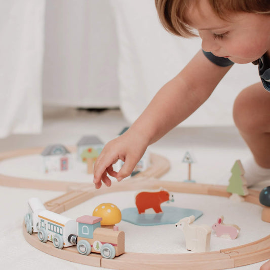 A young child gently placing a train carriage on the wooden track, surrounded by colourful woodland animals and scenery.
