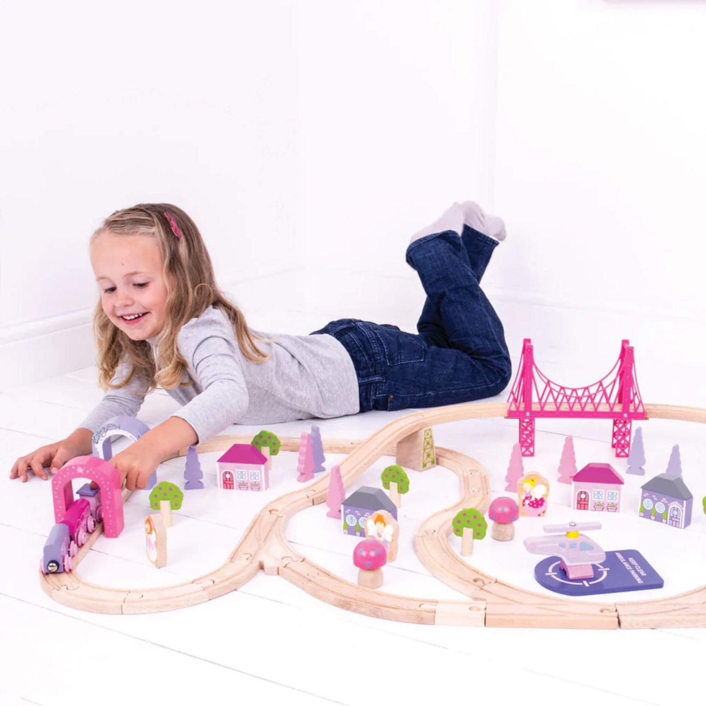 Child playing with the Bigjigs Fairy Town Train Set, guiding a pink train through a pastel tunnel surrounded by fairy figures, trees, and houses on a wooden floor.