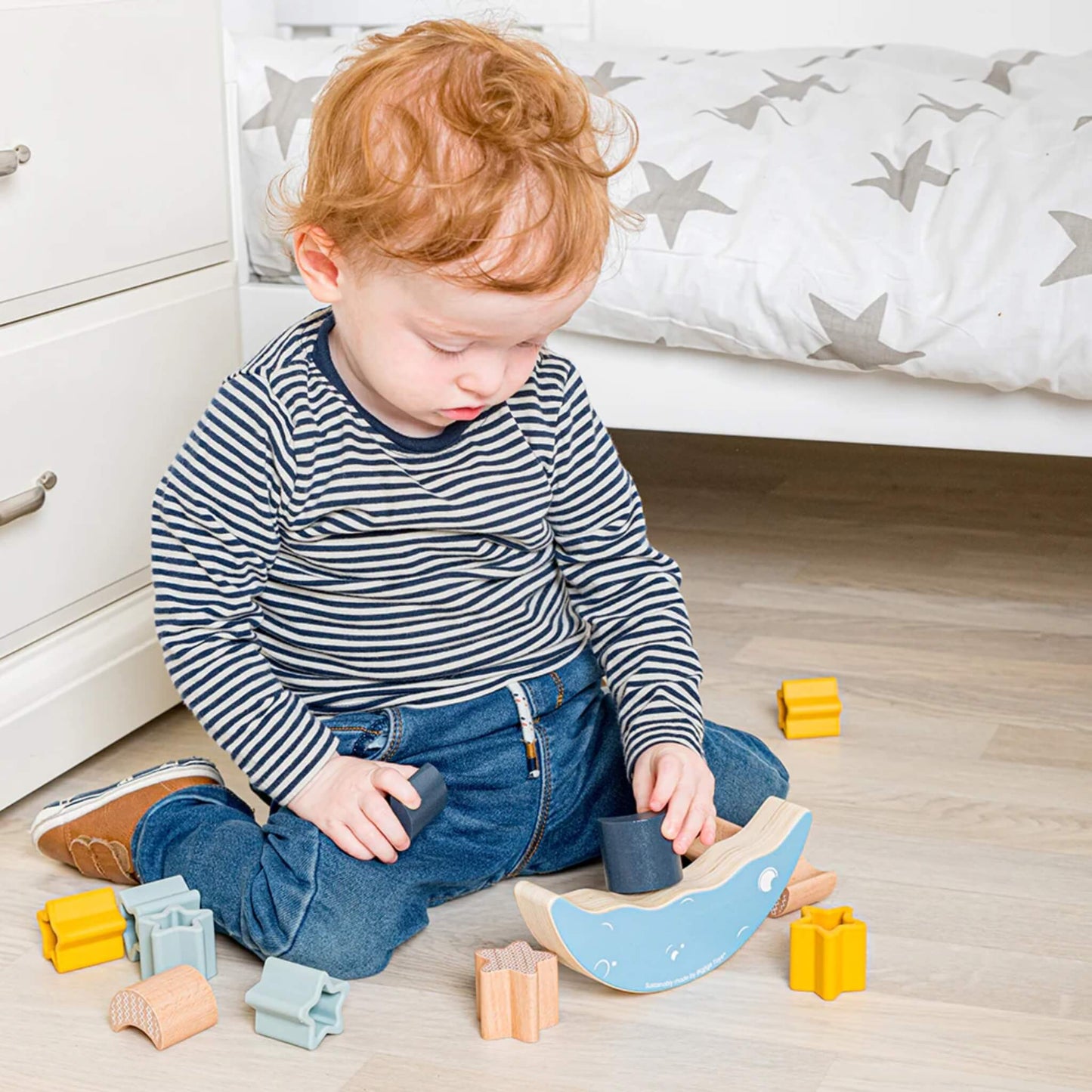 Toddler sitting on a bedroom floor, carefully placing pieces onto the Bigjigs Moon Balance Stacker, surrounded by colourful stars and moons.