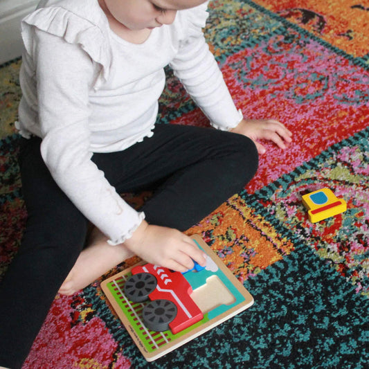 Young child carefully placing a chunky train piece into a wooden baseboard, with a bright and colourful train scene in the background.