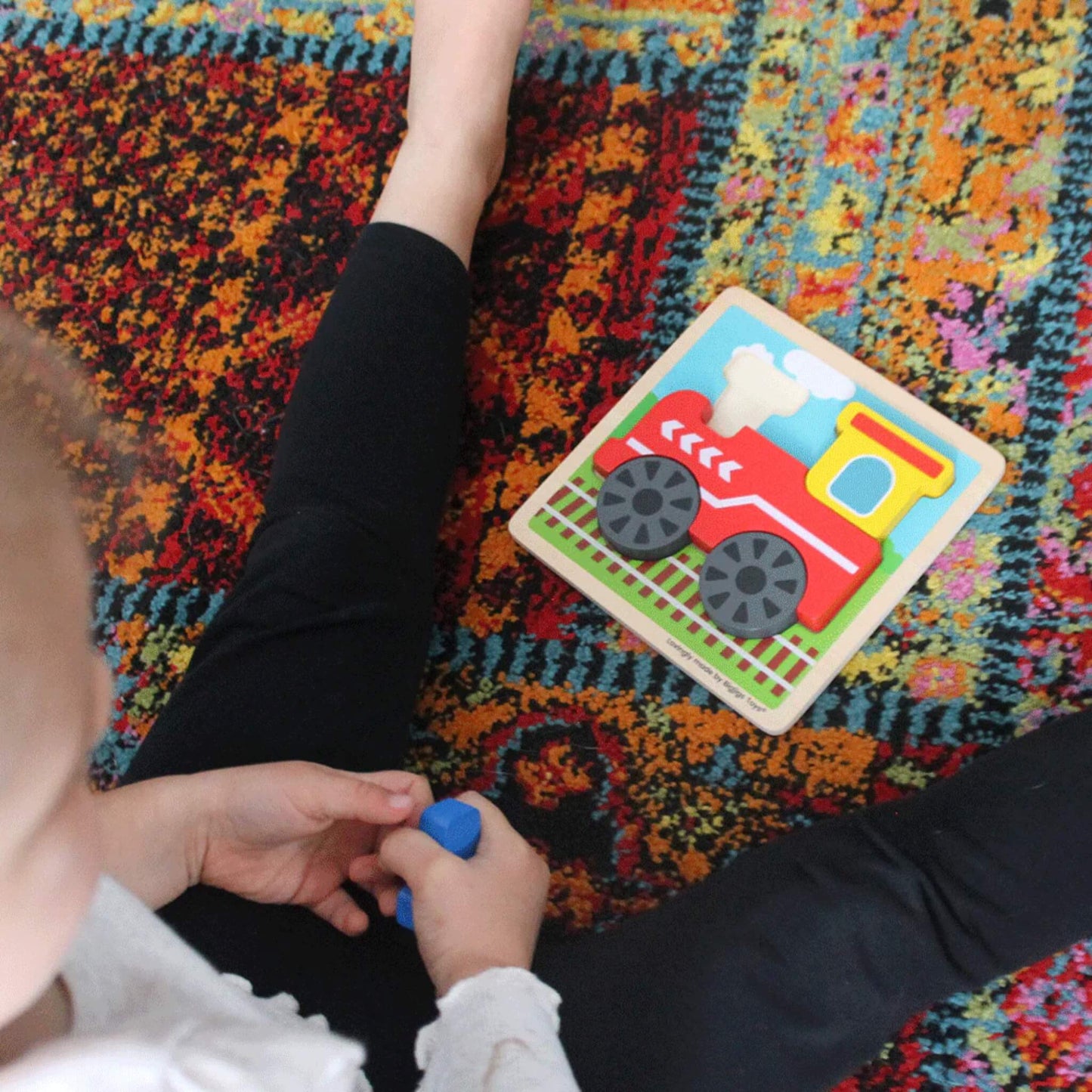 Child holding a blue puzzle piece while looking at a completed chunky train puzzle on a colourful patterned rug.