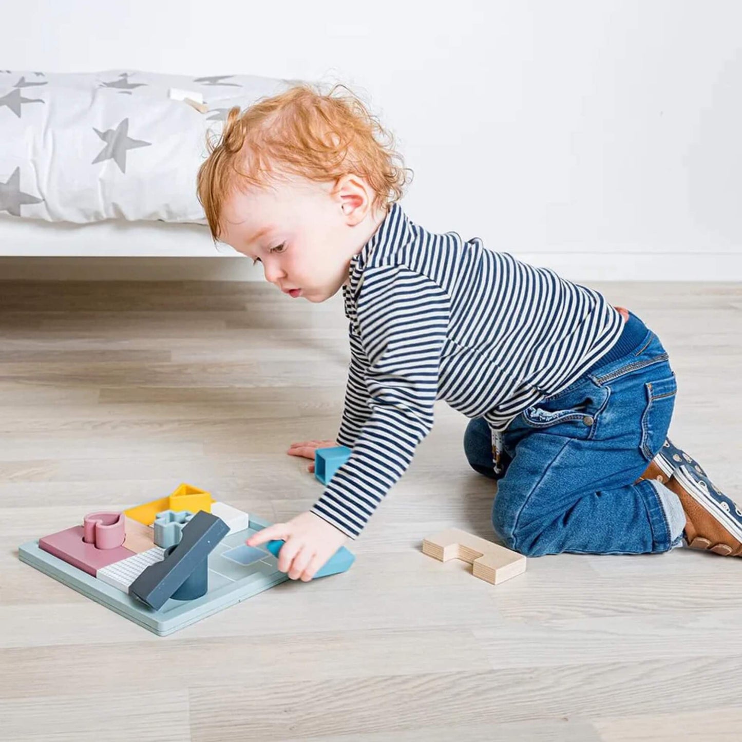 Toddler kneeling on the floor and reaching to place a wooden piece into the Bigjigs Toys Mosaic Puzzle, with other puzzle shapes scattered around.