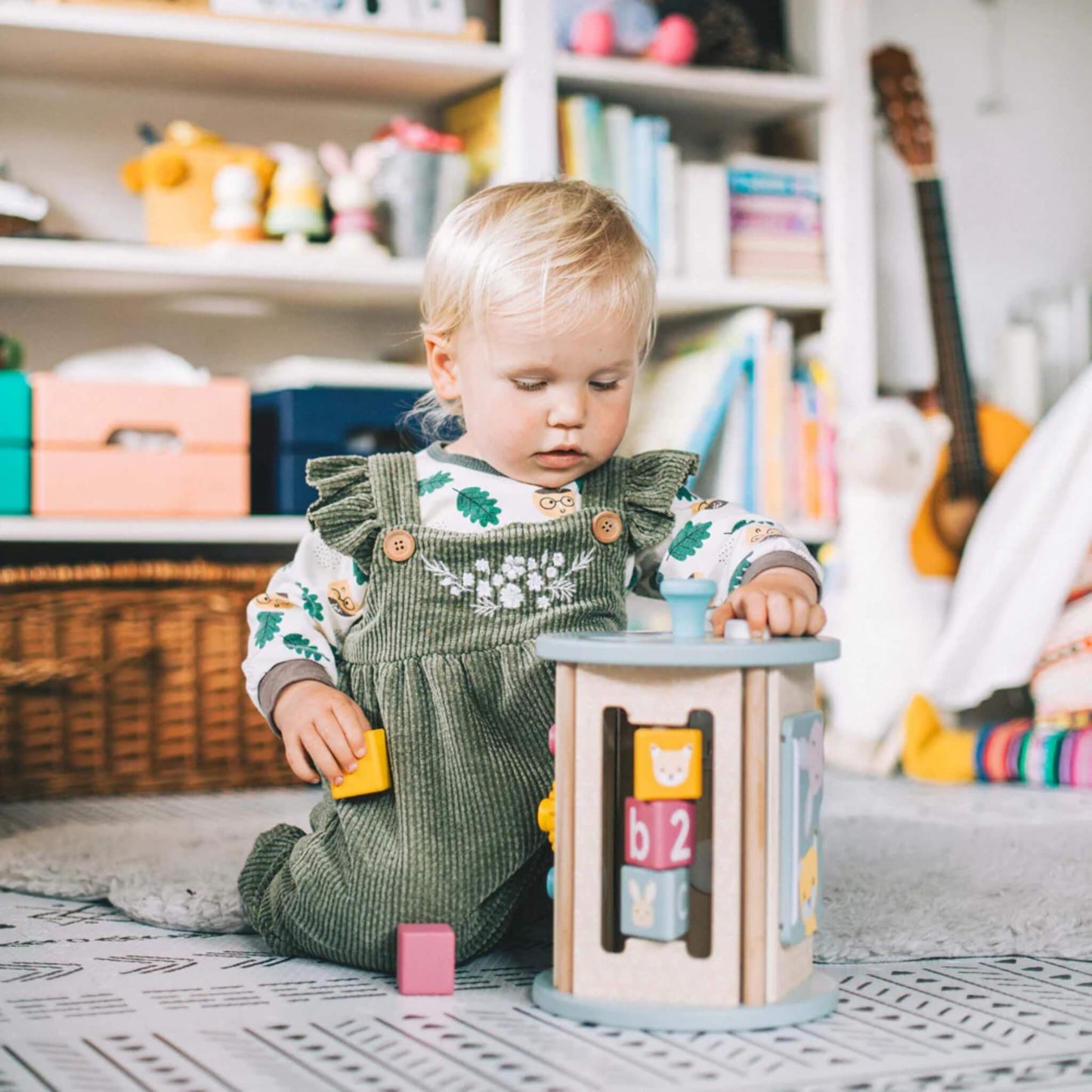 Child sitting on a soft rug, holding a yellow wooden block while interacting with the Bigjigs Rolling Activity Sorter in a warm, home play space.