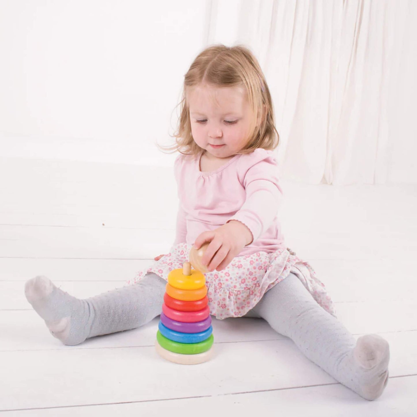 Toddler in a pink top and floral skirt placing the final piece on a fully assembled rainbow stacker.