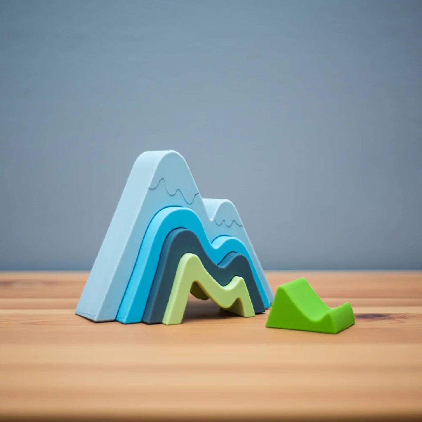 Toy silicone Bigjigs Mountain Stacker fully built on a wooden surface with soft lighting, featuring a mountain silhouette in gentle, natural colours.