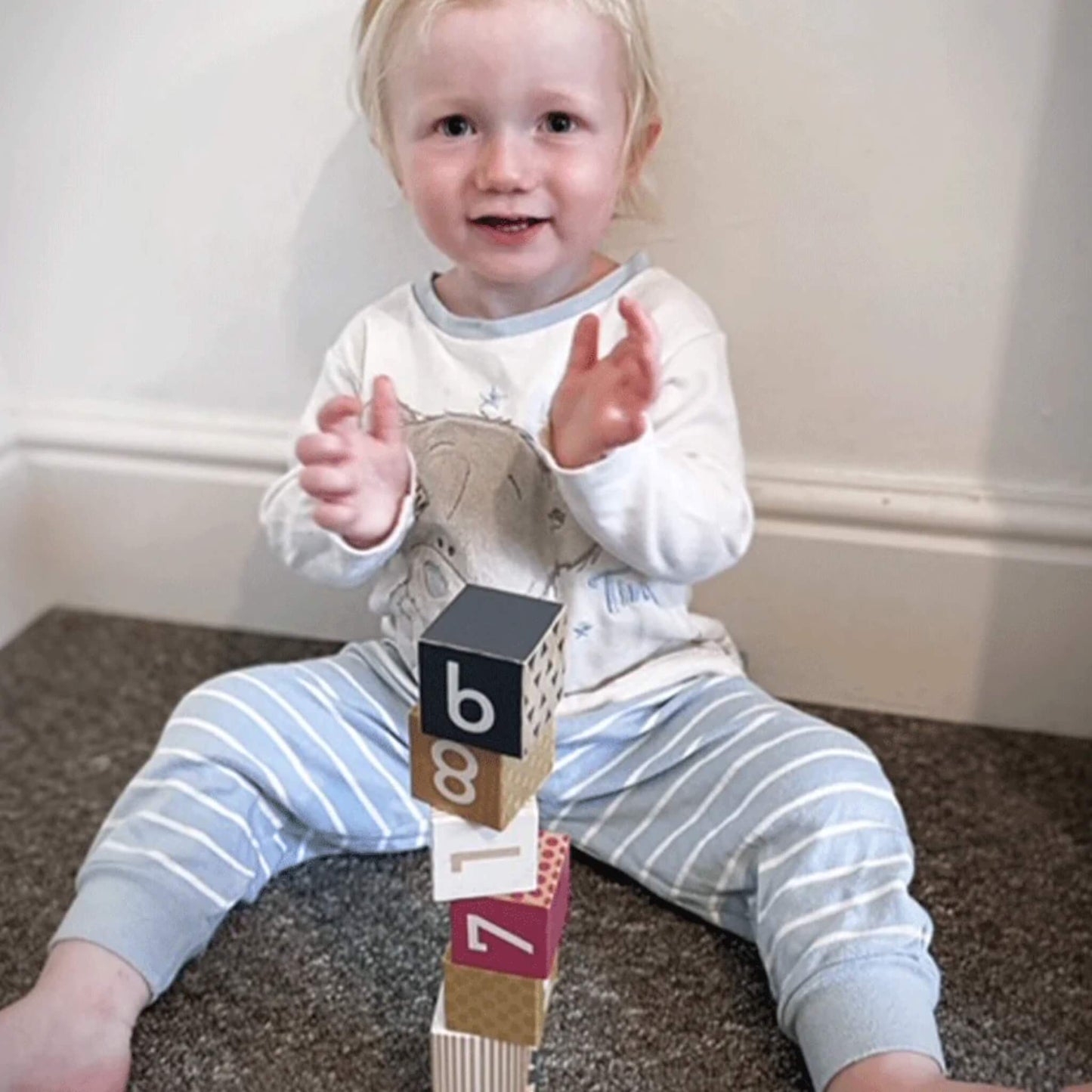 Toddler smiling and clapping while sitting on the floor with Bigjigs Toys Wooden Number Blocks stacked in a tall tower beside them.