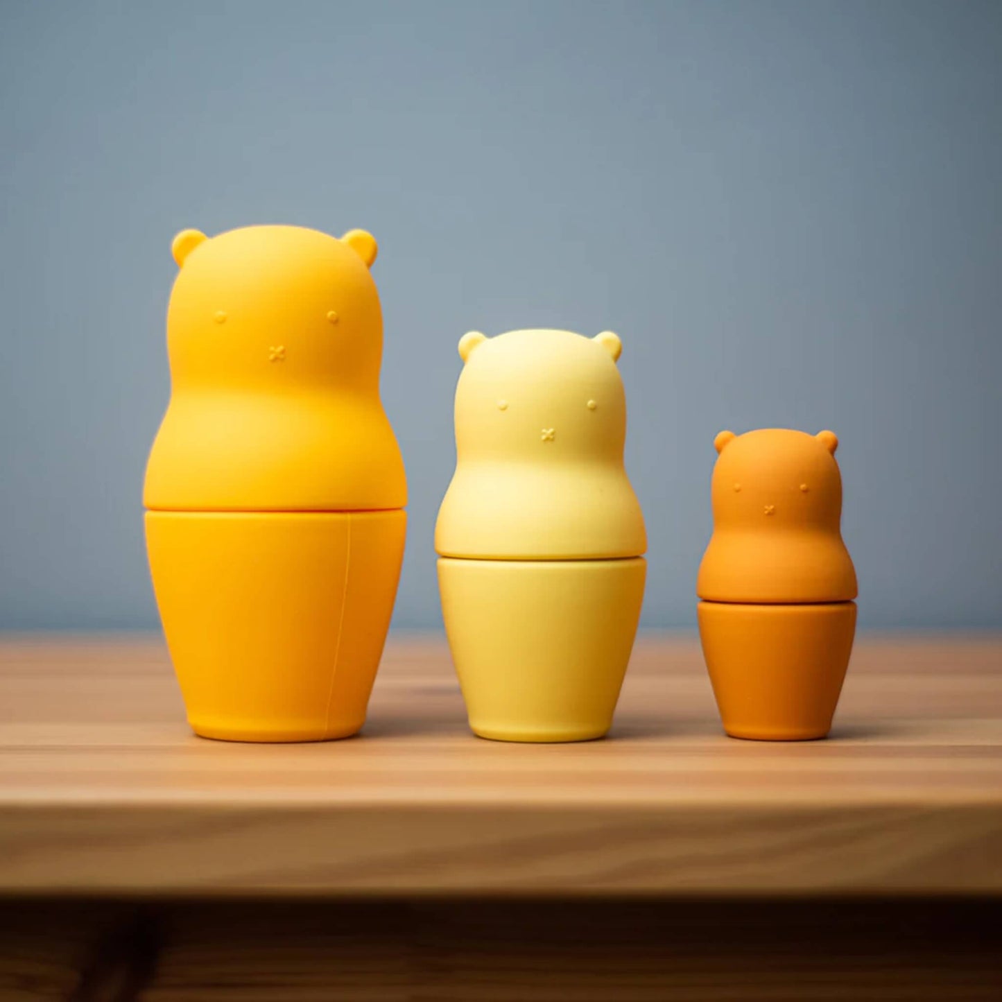 Three Bigjigs Silicone Nesting Bears lined up on a wooden surface with a soft blue wall background, showing full set upright.