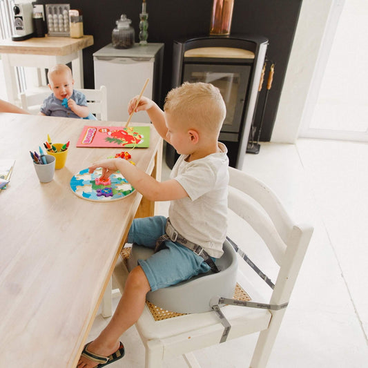 Toddler using the Bumbo Booster Seat in Cool Grey while seated at a dining table, painting a puzzle.
