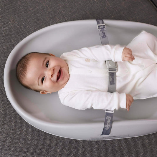 Smiling baby lying on the Bumbo Changing Pad in Cool Grey, safely secured with the 3-point strap on a dark carpeted floor.