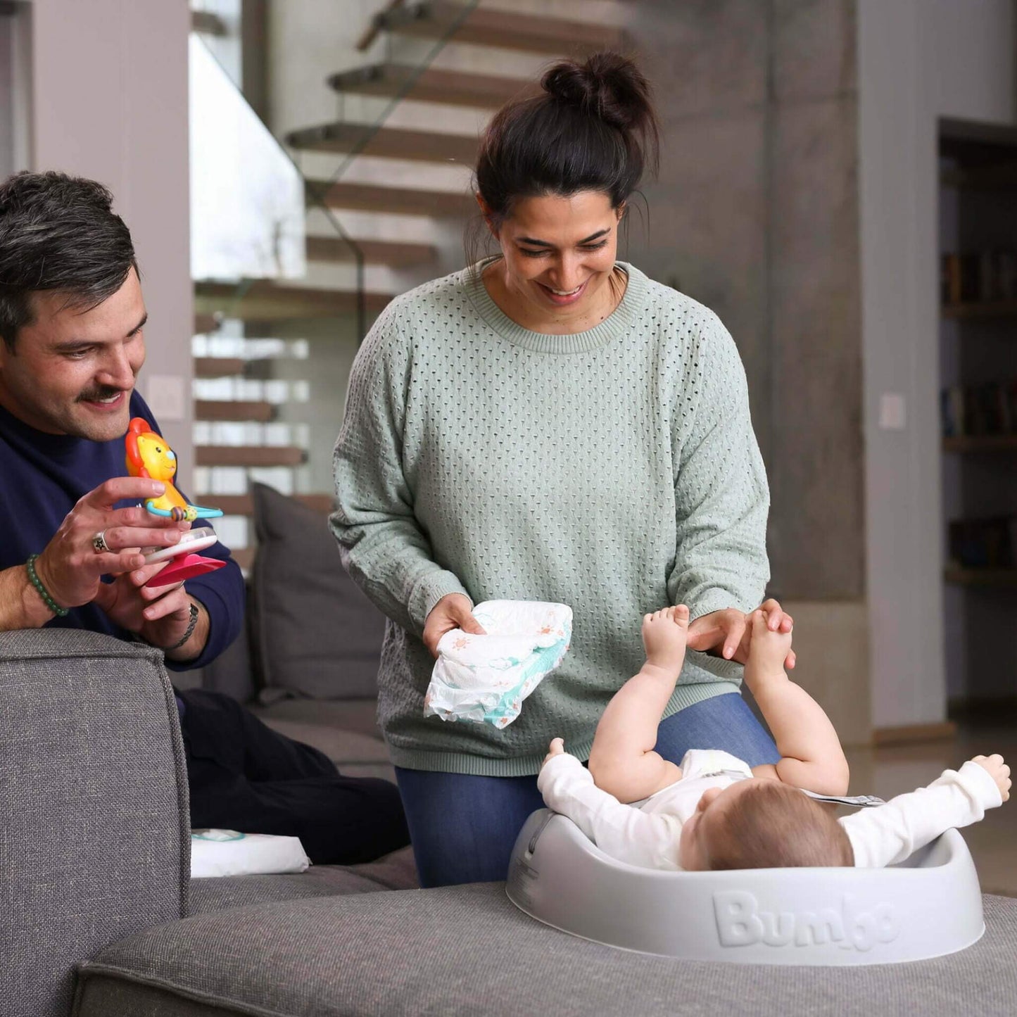 Mother changing a baby on the Bumbo Changing Pad while smiling father holds a toy, all seated together in a cosy modern living space.