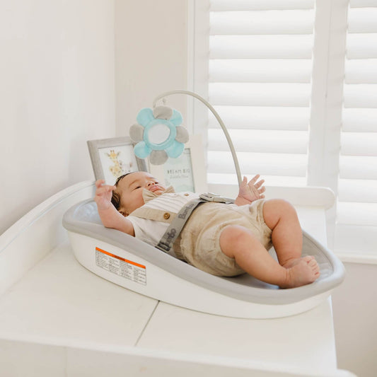 Baby lying on the Bumbo Max Changing Pad in Cool Grey, secured with the safety harness and reaching for a plush toy mobile.