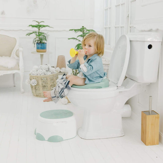 Toddler sits on a Bumbo toilet trainer in light green on a toilet, playing with a yellow trumpet, with a step stool nearby.
