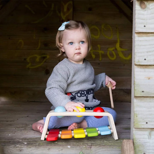 Toddler sitting on wooden decking holding the toy and yellow ball, with the Pound & Tap Bench placed on her lap.