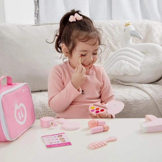 Young girl smiling while pretending to apply wooden toy makeup from a pink beauty set, with play accessories neatly spread out on the table.