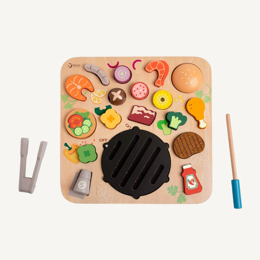 Wooden BBQ play board with removable food pieces including burger bun, prawn, corn, broccoli and steak arranged around a central grill.