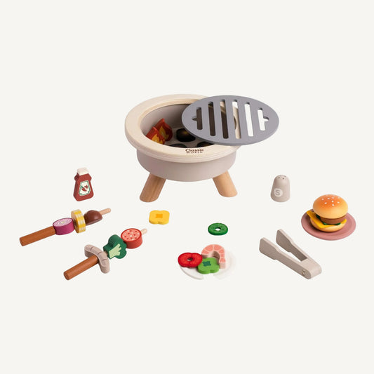 Wooden BBQ toy set with grill opened to reveal play coals, surrounded by food pieces, skewers, tongs and condiments on a plain background.