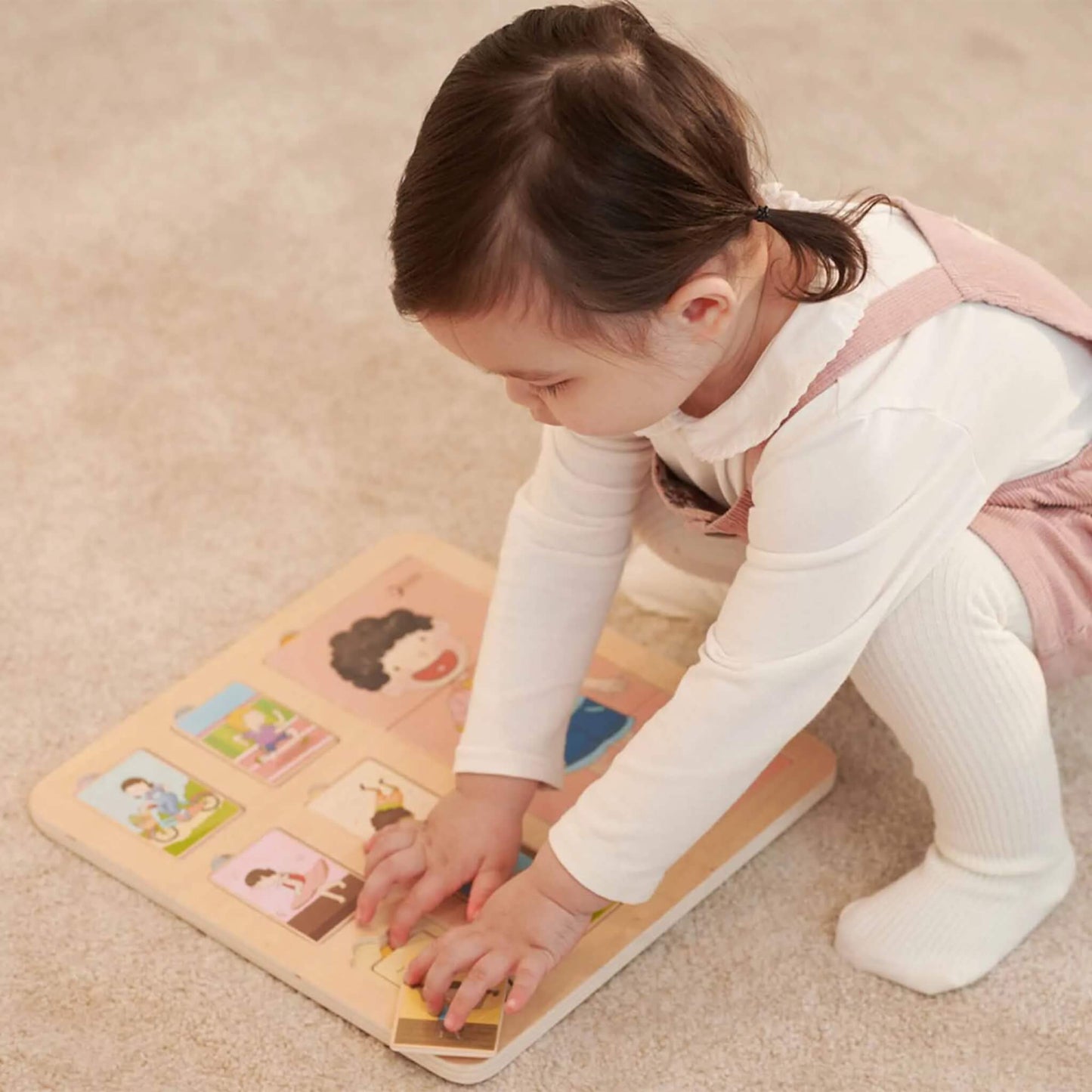 Toddler in white and pink clothing crouched over a wooden puzzle, carefully arranging pieces on the board.