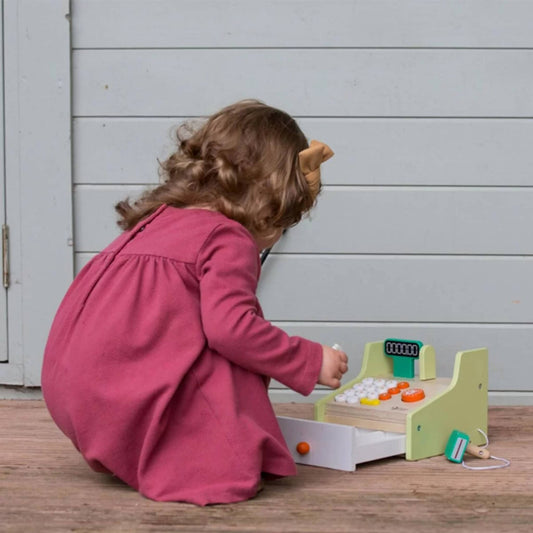 Toddler in a pink dress squatting on wooden floor, playing with the Classic World cash register, focused on pressing buttons with drawer open and toy money nearby.
