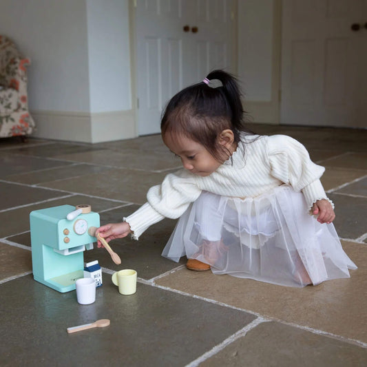 Toddler in a white dress crouching on a stone floor, reaching out to play with the wooden coffee maker set and accessories.