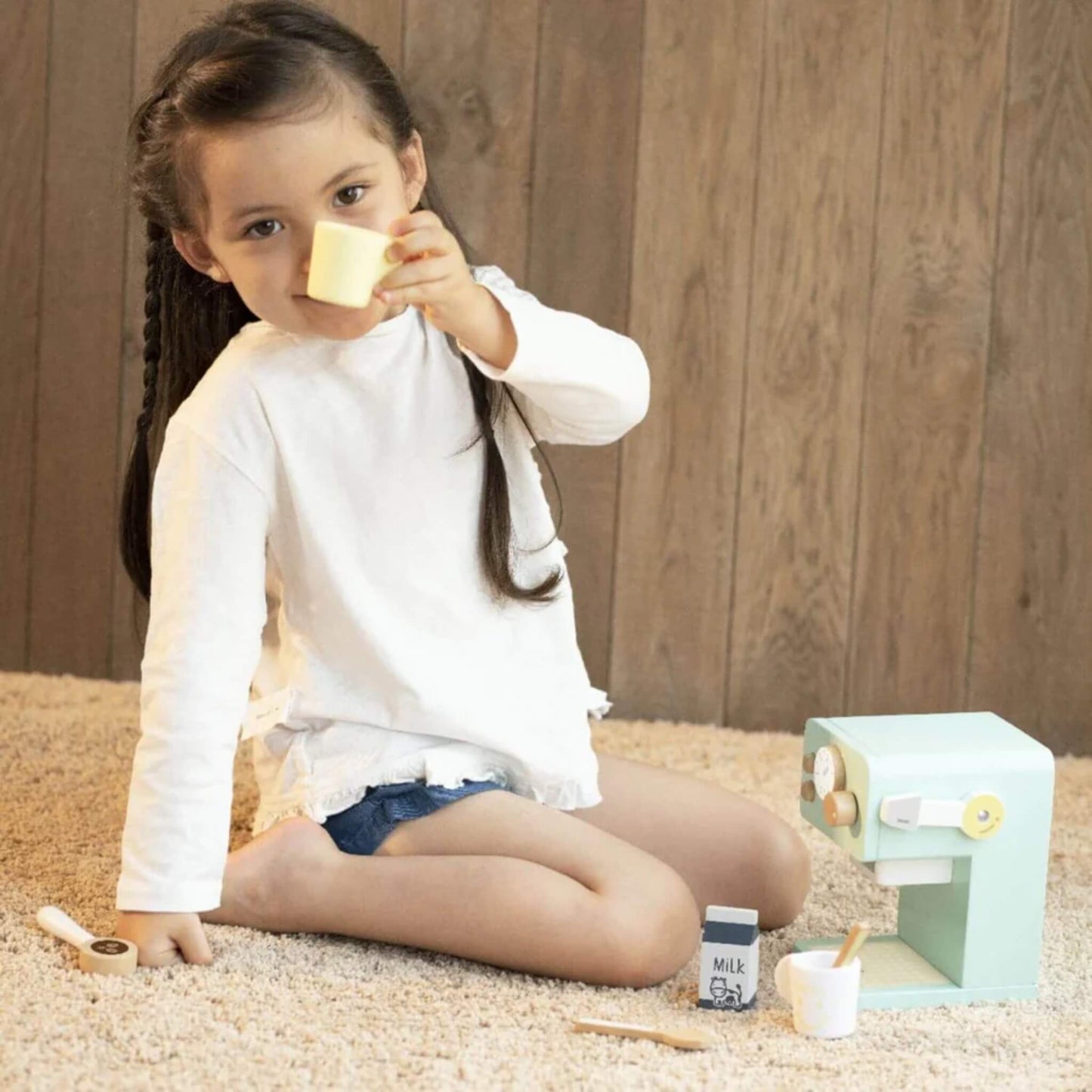 Young child sitting on a soft carpet, lifting a yellow mug in a playful toast next to the mint green wooden coffee machine.