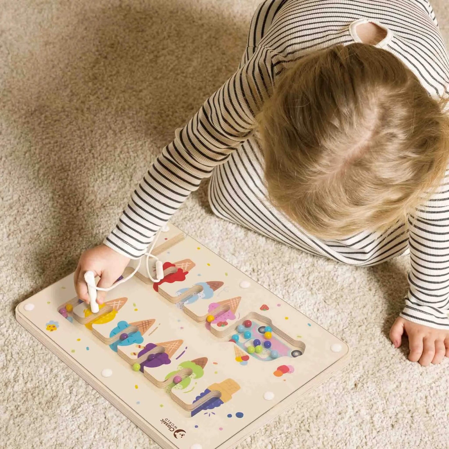 Child sitting on a cream carpet, using a magnetic pen to move coloured balls through a wooden maze with ice cream illustrations.