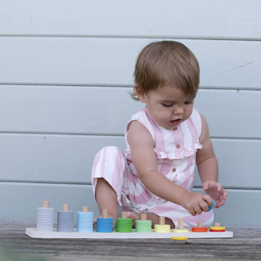 A toddler in a pink and white striped romper sitting on wooden decking, focused on stacking blue discs onto the peg of a colourful counting toy with numbered slots.