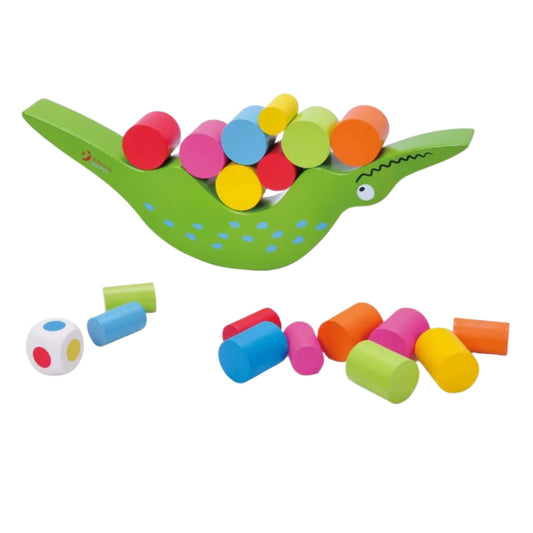 Green crocodile-shaped wooden balance toy with colourful cylinders spilling around it on a white surface, alongside a large white wooden die showing red, green, and blue dots.