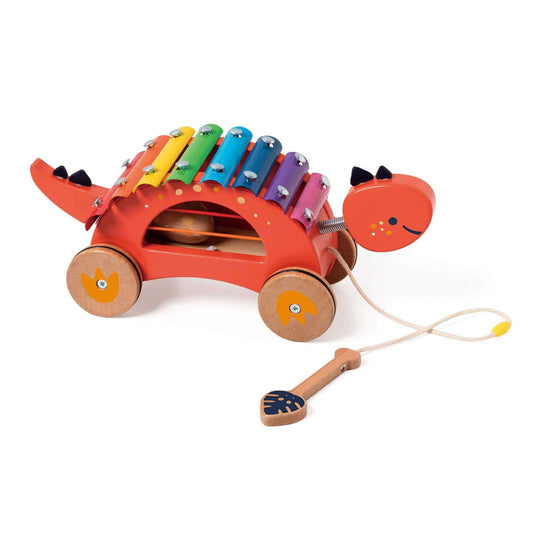 Side view of a red dinosaur-shaped pull-along xylophone with rainbow metal keys, wooden wheels, pull cord, and included mallet placed on the floor.