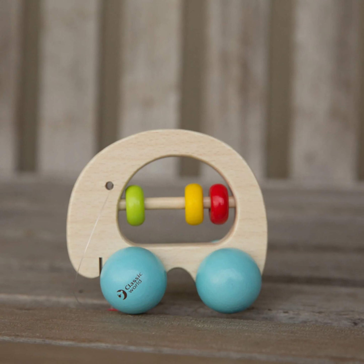 A wooden elephant-shaped rattle with blue wheels and three colourful beads on a central rod, resting on a wooden surface against a blurred neutral background.
