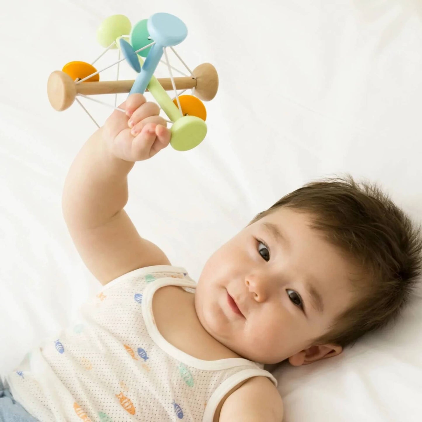 Baby lying on a white bed, smiling and holding up a colourful wooden sensory ball with both hands.