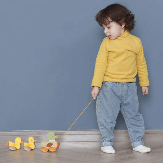A toddler wearing a yellow top and blue jeans holding a string and pulling a wooden duck family toy across a wooden floor.