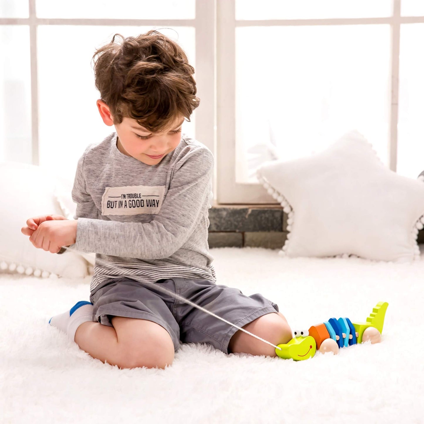 Child sitting on a soft white rug, gently pulling a string-attached wooden crocodile toy while smiling and looking down.