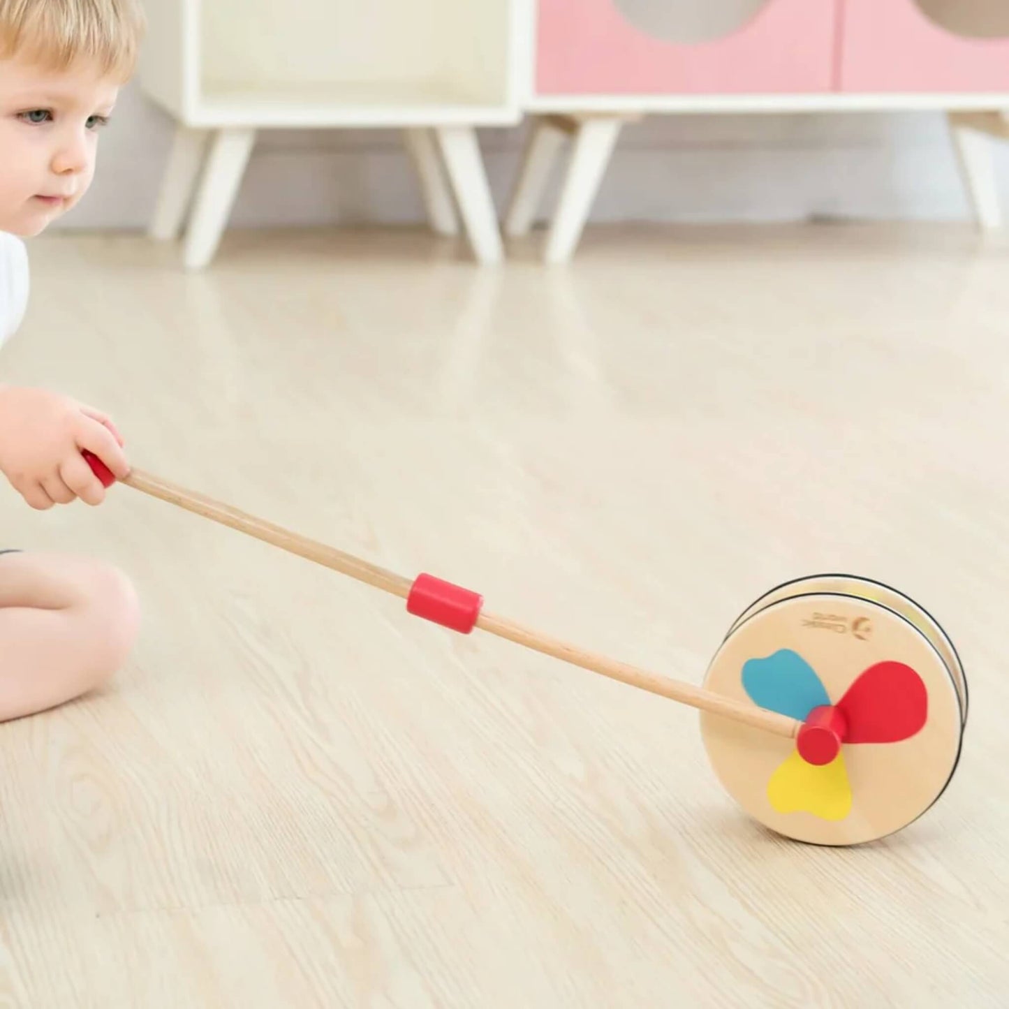 Child crouching indoors, gently rolling a wooden push toy forward, with red, yellow, and blue petal shapes on the wheel face.