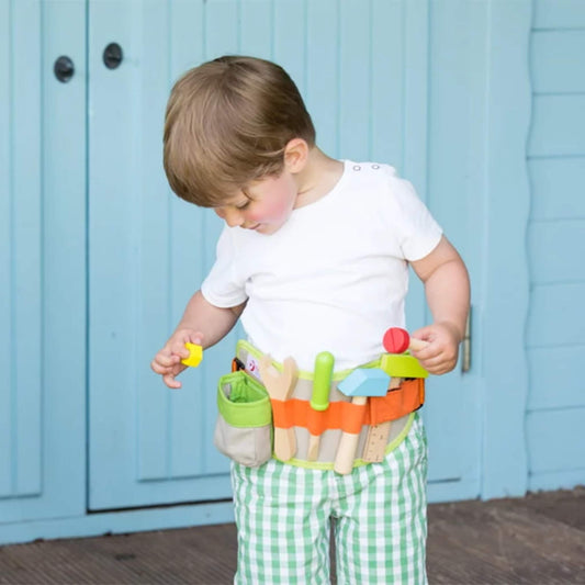 A toddler wearing a colourful tool belt with wooden tools, looking down at the belt while holding a yellow block and standing in front of blue wooden doors.
