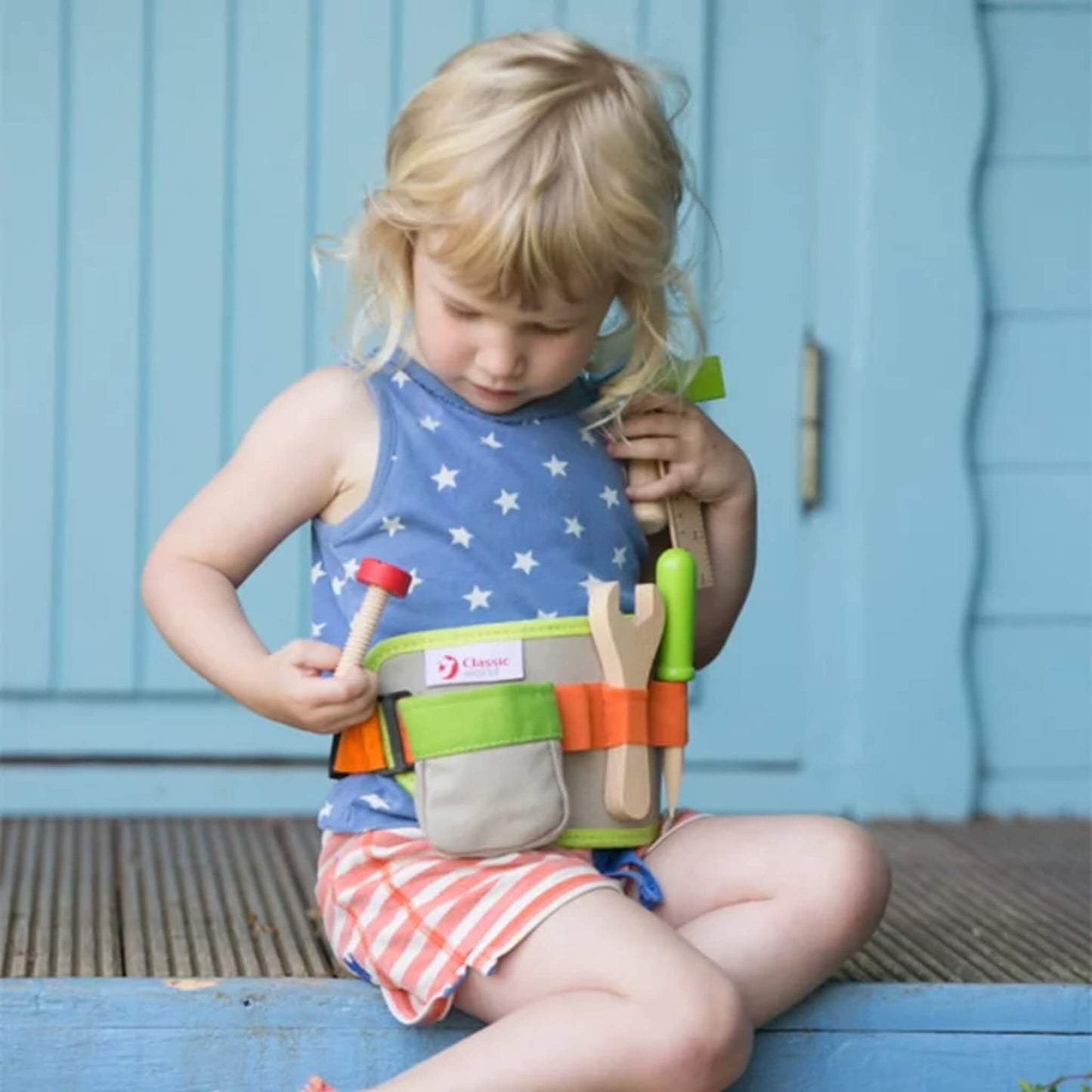 A young girl sits on a wooden step, exploring the items inside her toy tool belt which includes wooden tools and a fabric pouch with the Classic World logo.