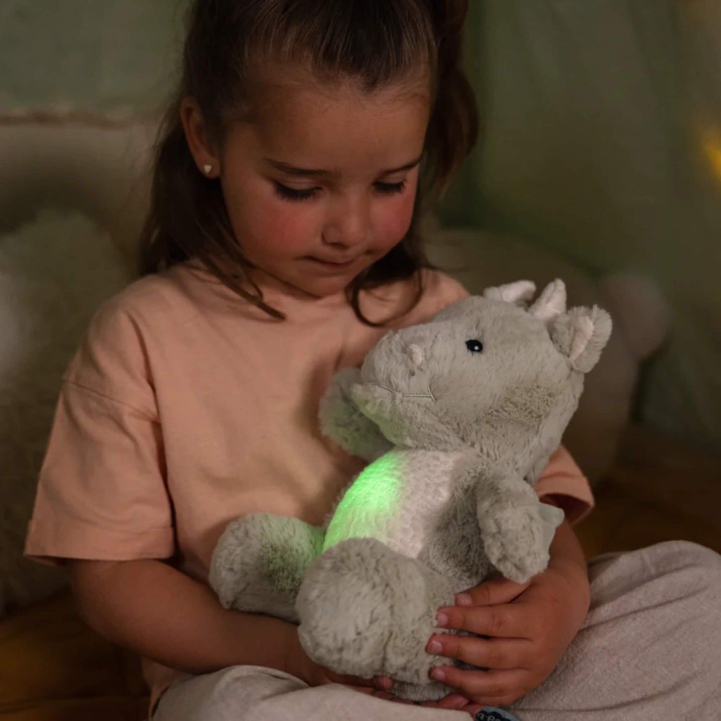 A girl in a peach top holds a plush dragon toy on her lap, watching as its tummy glows green in the dim light of a cosy room.