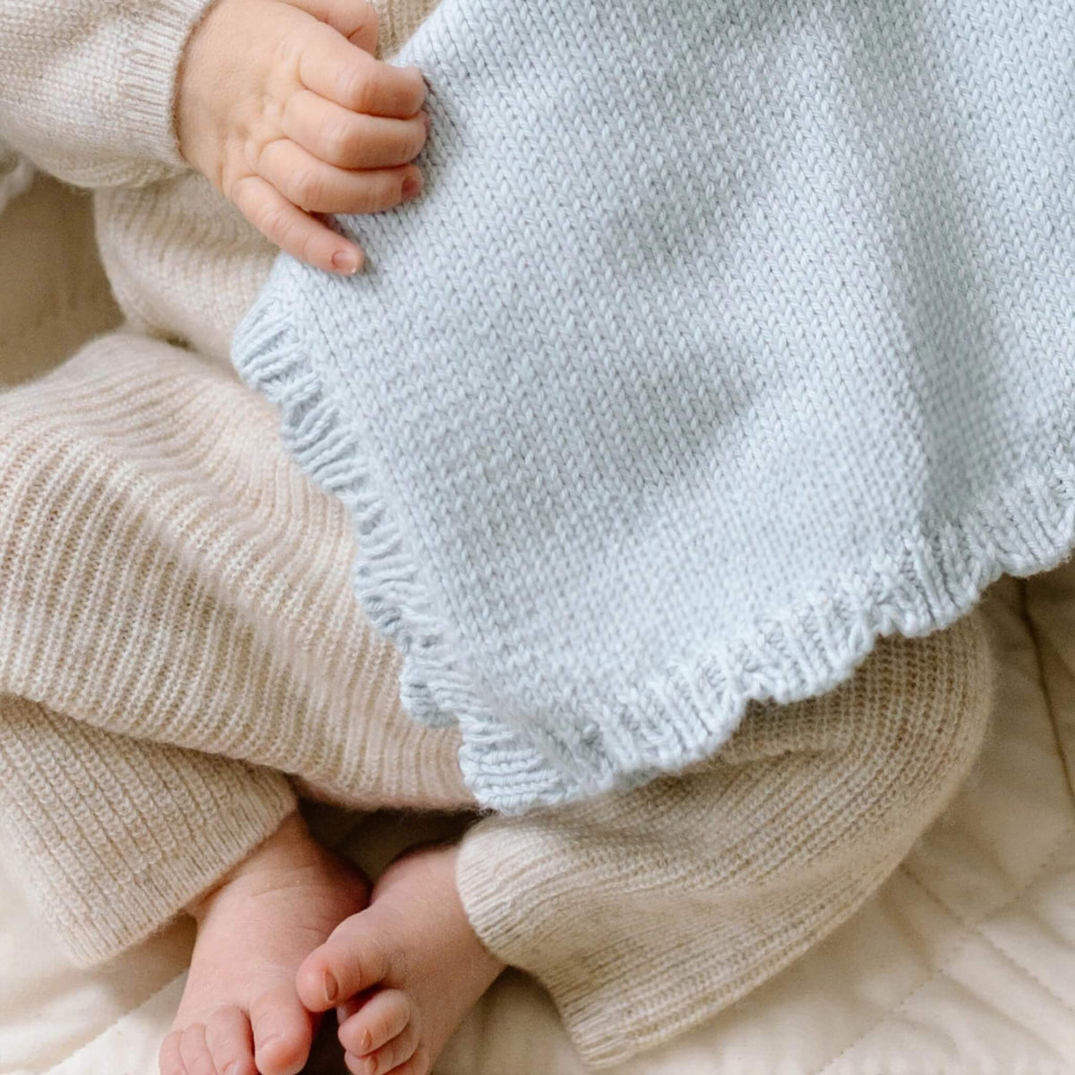 Newborn baby's hand holding a pale blue knitted comforter, showing the soft organic cotton fabric and scalloped edging.
