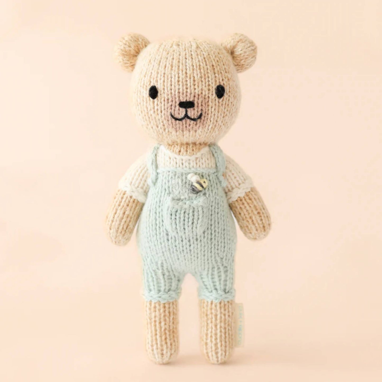 A hand-knit cotton doll standing upright against a neutral background, wearing pale green overalls with a small stitched detail on the chest.