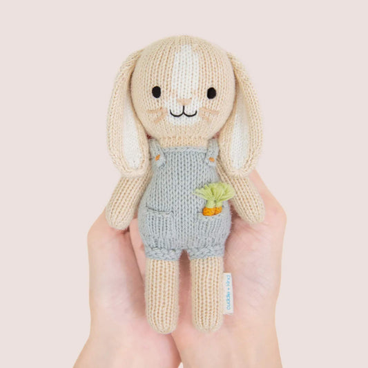Small knitted bunny doll held in hands showing size, wearing soft grey overalls with a tiny carrot detail in the pocket.