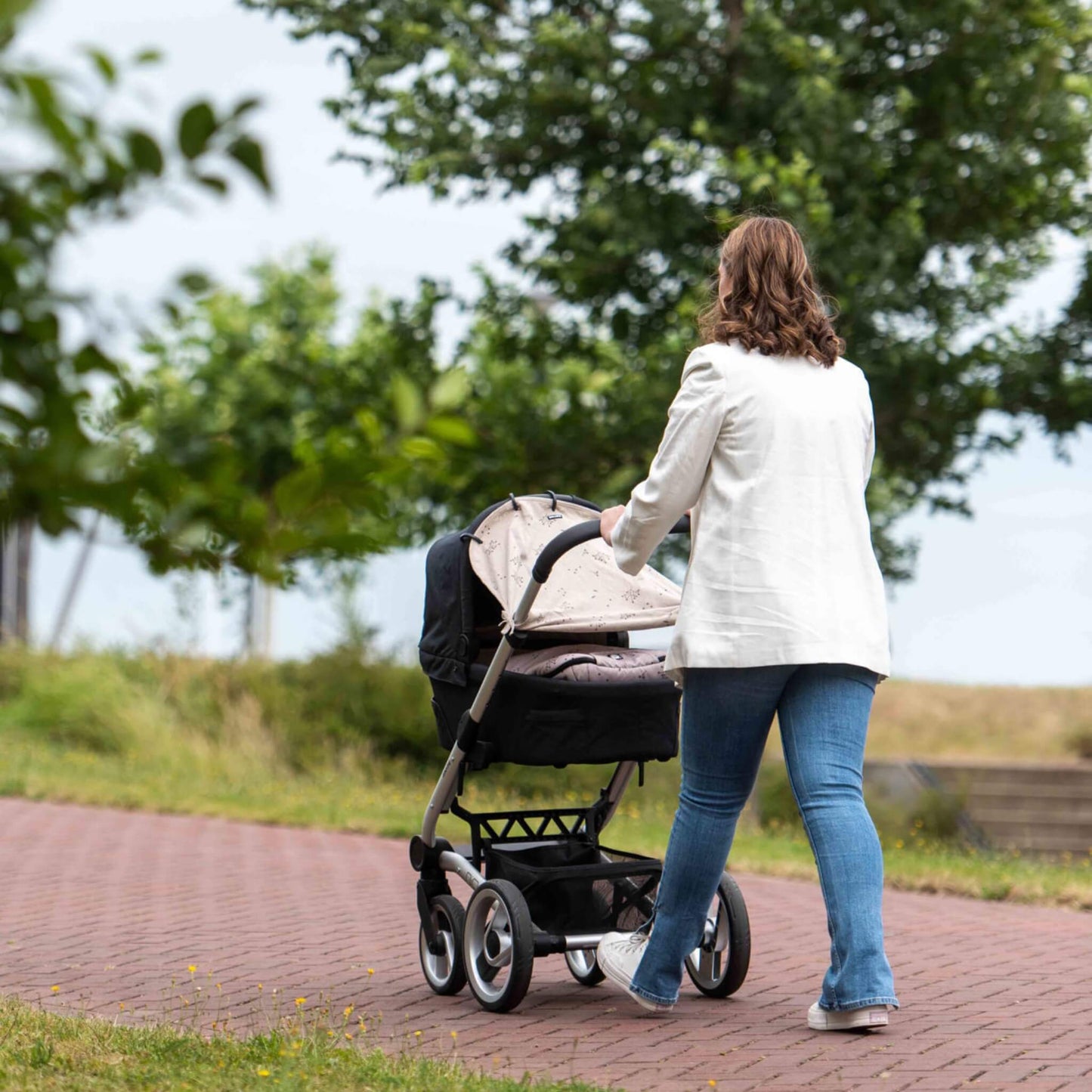 A woman pushing a pram fitted with a beige botanical print sunshade while walking outdoors on a sunny day.