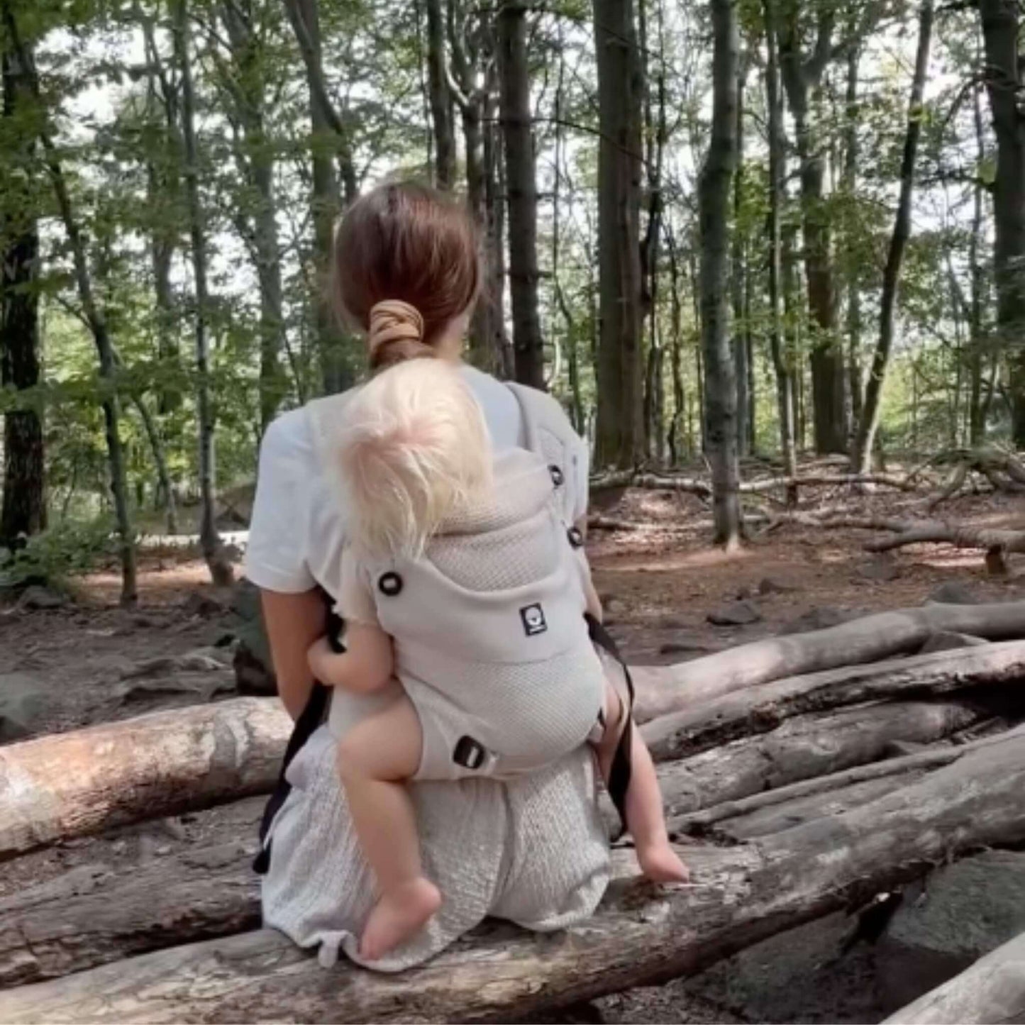 A baby carrier with a baby worn on a woman's back while she sits on a fallen log in a forest.