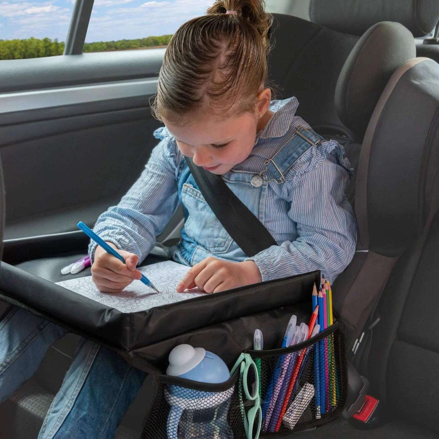 A young child sitting in a car seat using a black travel table to colour in a book, with mesh side pockets holding pencils, a water bottle, and sunglasses.