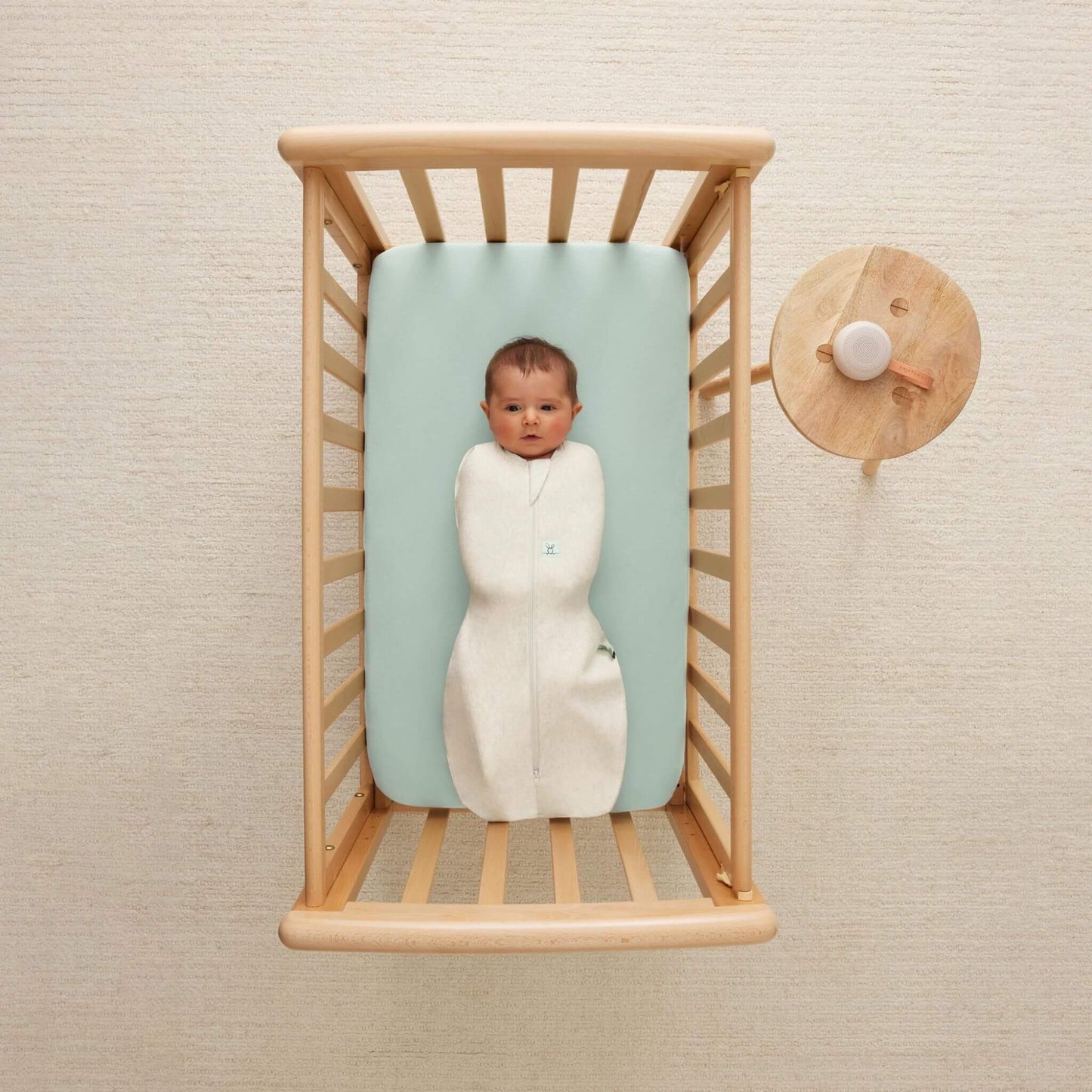 Baby lying securely in a wooden cot, wrapped in a Swaddle Bag in oatmeal, on a pastel blue mattress.