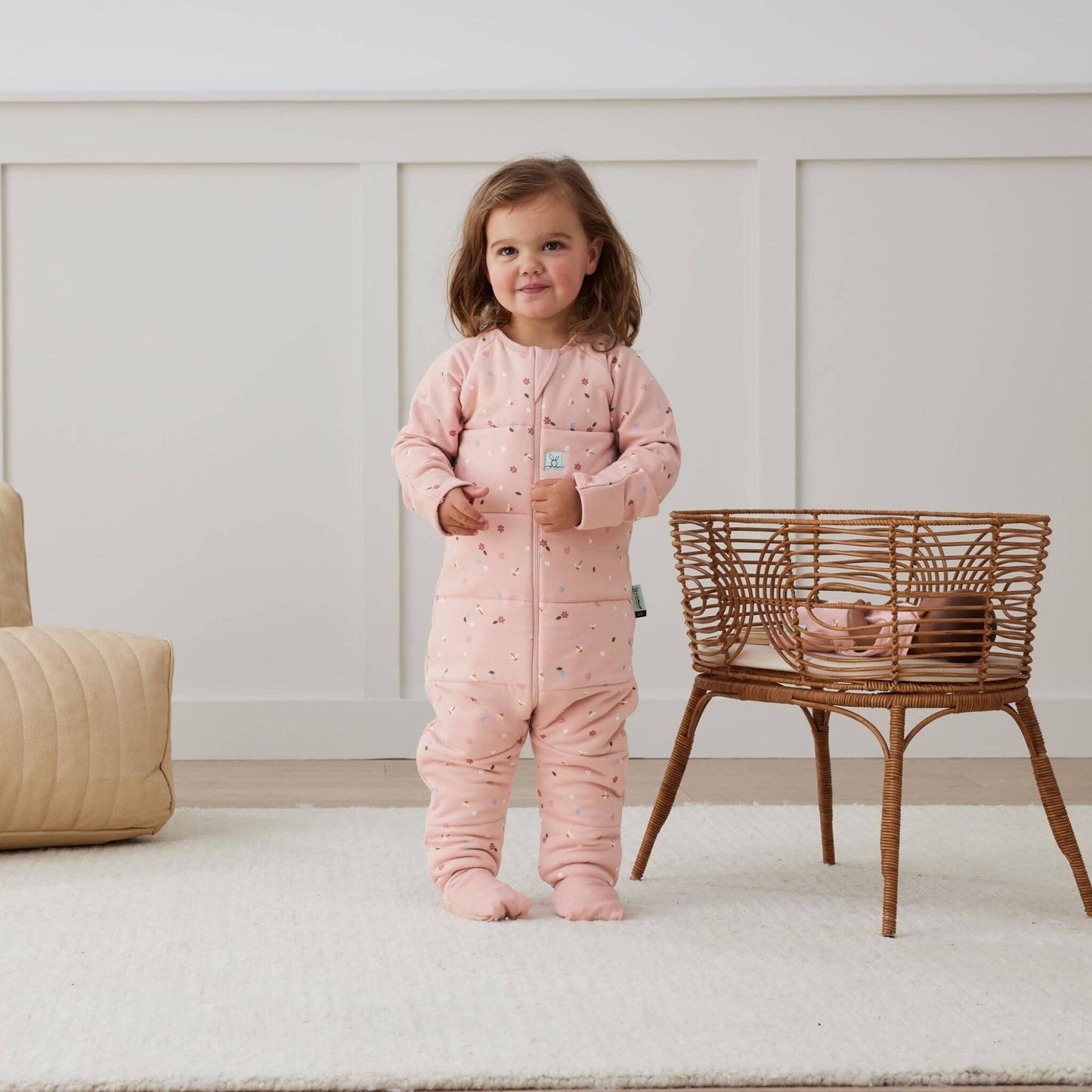 A toddler stands in a winter weight Sleep Onesie with a Daisies print, featuring a soft pink design with delicate floral details. Perfect for cosy sleep, the Onesie offers a snug, breathable fit with fold-over foot cuffs and a three-way zip for added convenience.