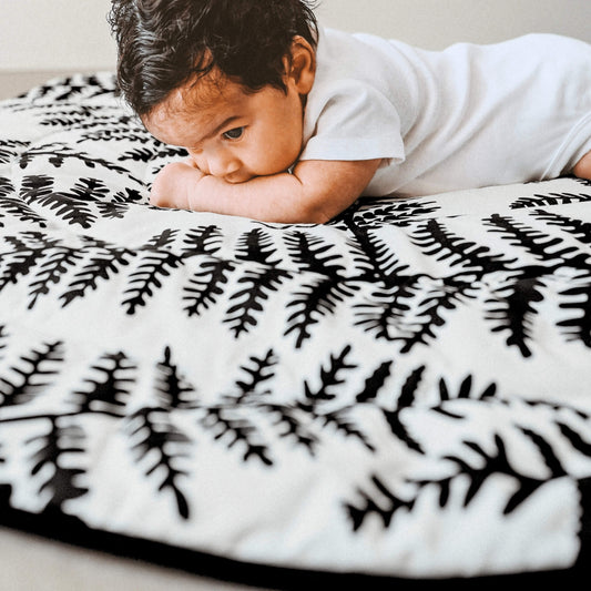 Baby lying on a padded playmat with high-contrast black fern pattern during tummy time, focusing closely on surface.