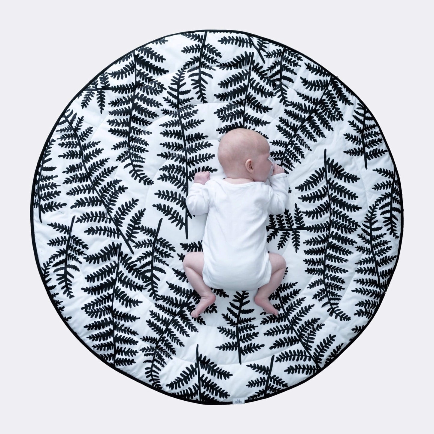 Overhead view of baby lying centred on large round cushioned playmat with bold black botanical print for tummy time.